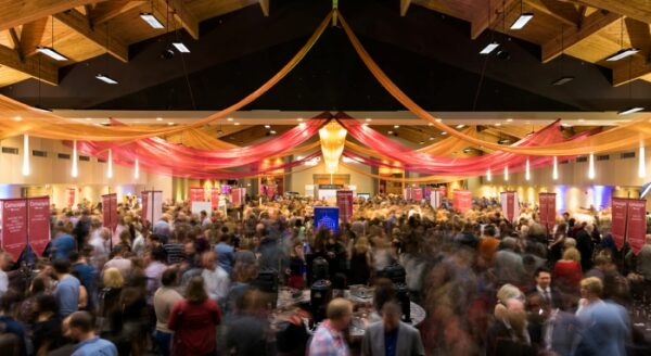 A lively indoor event featuring a large crowd of people gathered under colorful drapery. The scene shows vibrant decorations and an energetic atmosphere, indicating a social gathering or celebration.