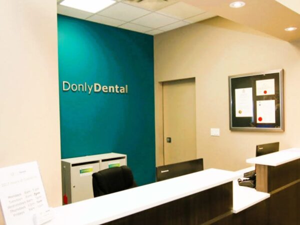 A dental office reception area featuring a modern design with a desk, a teal accent wall displaying the name "DonlyDental," and framed information on the wall.