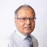 A middle-aged man with glasses and short, grey hair is smiling, wearing a checkered shirt and tie, against a plain white background.