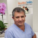 A medical professional wearing a blue scrub top smiles in an office setting. In the background, there are decorative plants and a sign for "Bloor West Smiles," indicating a focus on dental care.