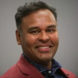 A smiling man with short, dark hair wearing a red blazer over a blue patterned shirt. He has a friendly expression against a neutral background.