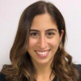 A woman with long, wavy brown hair smiles at the camera. She is wearing hoop earrings and a black top, set against a plain light background.