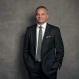 A professional man in a dark suit and tie stands confidently with his hands in his pockets, smiling softly against a neutral background.