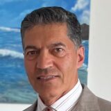 A man with dark hair and a confident smile stands in front of a colorful ocean mural, wearing a light suit and patterned tie.