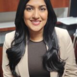 A confident individual with long black hair is smiling, wearing a light-colored blazer over a dark top, seated in an office environment with wooden cabinetry in the background.