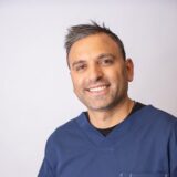 A smiling individual wearing a blue medical scrubs top poses against a plain background, conveying a friendly and approachable demeanor.