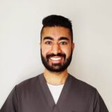 A smiling man with a beard is wearing scrubs, standing against a plain background.