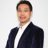 A man in a dark blazer and white shirt smiles confidently against a plain white background.