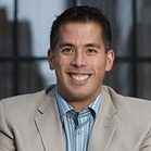 A man is smiling while wearing a light-colored suit jacket over a blue striped shirt. The background features blurred architectural elements, suggesting an urban setting.