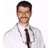 A smiling male dentist wearing glasses, a white lab coat, and a tie, with dental tools around his neck, stands against a plain white background.