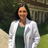 A young woman wearing a white lab coat stands outdoors, smiling. She is in front of a brick building, surrounded by greenery. The image conveys a sense of professionalism and youthfulness.