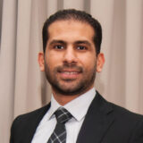 A professional man in a suit and tie is smiling for the camera, set against a soft, neutral background. His friendly demeanor conveys confidence and approachability.
