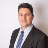 A professionally dressed man with short dark hair smiles confidently. He wears a black suit and a light blue shirt with a patterned tie, standing against a plain white background.