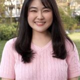 A young woman with long black hair smiles warmly while wearing a pink cable-knit sweater, standing outdoors in a green park setting with trees in the background.