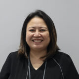 A smiling woman with medium-length dark hair is wearing a black top. She is facing the camera against a plain light gray background, conveying a friendly and welcoming demeanor.