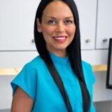 A smiling woman with long black hair wearing a turquoise blouse stands in an office environment, featuring light-colored cabinetry in the background.