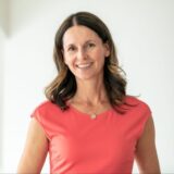 A smiling woman with long, wavy hair and wearing a coral-colored top stands in a bright, minimalistic setting. She exudes a cheerful and approachable demeanor.