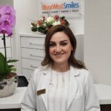 A smiling professional woman in a white coat stands in a dental office, with a floral arrangement and a sign reading "Bloor West Smiles" in the background.