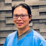 A smiling individual wearing blue scrubs and glasses stands in front of a textured gray wall.