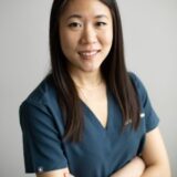 A woman dressed in medical scrubs smiles confidently while standing with her arms crossed against a neutral background.