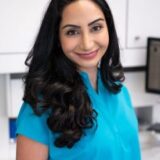 A smiling woman with long, dark hair wears a bright turquoise blouse. She stands in a well-lit, modern office environment, appearing confident and approachable.