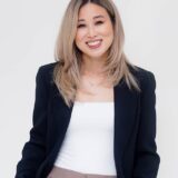 A woman with long, light brown hair smiles confidently while wearing a black blazer over a white top. She stands against a plain white background, exuding a professional and approachable demeanor.