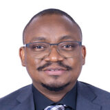 A smiling man with glasses, wearing a black suit and a patterned tie, poses for a professional headshot against a plain white background.