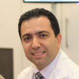 A smiling healthcare professional in a white lab coat, sitting in an office setting. The background includes a digital display, suggesting a clinical environment.