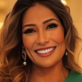 A woman with long, wavy hair smiles warmly, wearing elegant earrings and a green top. Her bright smile and confident expression convey a sense of joy and approachability. The background is softly blurred, emphasizing her features.