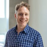 A smiling man with curly hair stands in a bright setting, wearing a blue and white checkered shirt. He appears cheerful and approachable, with a neutral background highlighting his expression.
