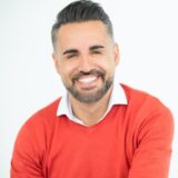 A man with a beard smiles warmly while wearing a red sweater over a collared shirt. He has short, neatly styled hair and is seated against a light background, conveying a friendly and approachable demeanor.
