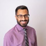 A smiling man with a beard and glasses is wearing a purple dress shirt and a striped tie. He is posed against a light background, exuding a professional and friendly demeanor.
