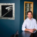 A man in a light shirt smiles while leaning on a reception desk in a modern office. Behind him is a bold artwork featuring a stylized dental instrument, set against a dark blue wall, with a glimpse of a glass-paneled door.