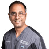 A smiling dentist in gray scrubs, standing confidently and looking directly at the camera. He has short, dark hair and glasses, representing a friendly and professional demeanor in a dental practice setting.