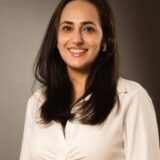 A smiling woman with long, dark hair is wearing a white blouse. The background is a soft gradient of gray, highlighting her cheerful demeanor and professional appearance.