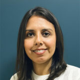A woman with dark hair and a warm smile is posing against a blue background, exuding a friendly and approachable demeanor.