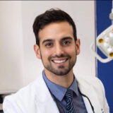 A smiling male dentist in a white coat stands confidently in a dental office, with bright lighting and dental equipment visible in the background. His friendly demeanor conveys approachability and professionalism.