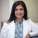 A smiling woman with long dark hair, wearing a white lab coat over a patterned shirt, sits casually in a modern office setting.