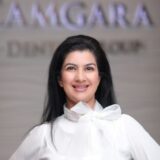A smiling woman with long black hair poses in a white blouse with a bow. The background features a sign that reads "Camgara Dental Group."