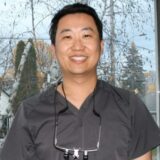 A smiling individual wearing scrubs and dental loupes stands in front of a window with a view of trees and a cloudy sky.