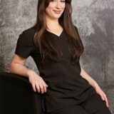 A young woman sits casually, wearing a black scrub uniform, with long, straight hair and a friendly smile. She poses confidently against a textured grey background, exuding a professional yet approachable demeanor.