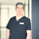 A smiling man wearing a black medical scrub stands in a well-lit environment. He has short, gray hair and is positioned near a doorway, conveying a professional and approachable demeanor.