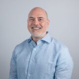 A smiling man with a bald head and a neatly trimmed beard is wearing a light blue button-up shirt. The background is plain and neutral.