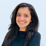 A young woman with long dark hair smiles warmly at the camera, wearing a black top against a soft blue background. Her friendly expression conveys a sense of approachability and positivity.