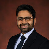 A smiling professional man wearing glasses and a formal suit, posed against a warm, gradient background. His expression exudes confidence and approachability.