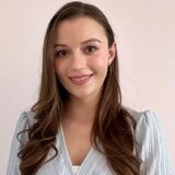 A young woman with long, wavy brown hair smiles at the camera, wearing a light blue blouse. The background is neutral, creating a warm and friendly atmosphere.