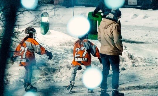 Two children in orange jerseys skate on a snowy rink, while an adult stands nearby. The scene is slightly blurred with glowing blue orbs in the foreground, suggesting a cold, winter evening atmosphere.