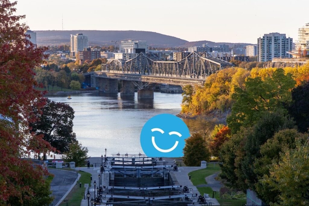 A scenic view of a river with a bridge in the background, surrounded by trees displaying autumn colors and city buildings on the horizon. A blue smiley face is overlayed on the image.