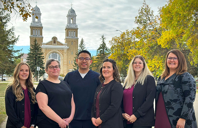 A group of six dental professionals, dressed in professional attire, stand outdoors with a historic building and trees in the background. The atmosphere is friendly, and the scene is brightened by the presence of the cheerful 123Dentist Smiley.