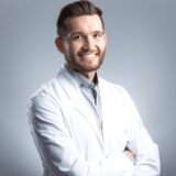 A smiling man in a white coat poses confidently with arms crossed, set against a light background. He appears friendly and professional, likely representing a healthcare or dental profession.